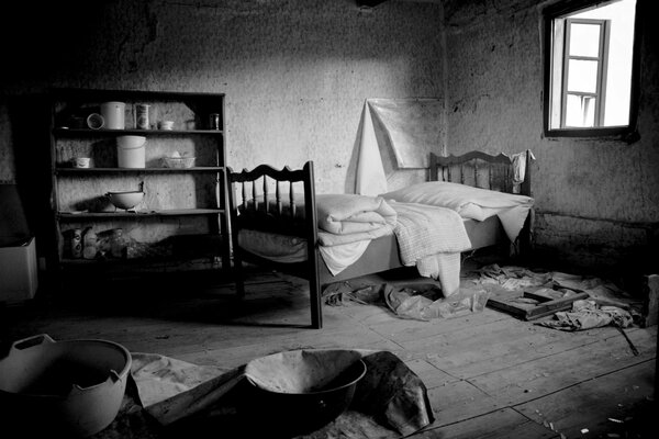 Une maison abandonnée par les gens avec des fenêtres ouvertes avec un lit non directionnel et des étagères avec des restes de vaisselle en noir et blanc