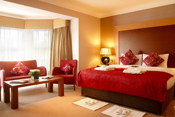 Nowoczesna sypialnia z podwójnym łóżkiem pod czerwoną narzutą i kącikiem do odpoczynku w oknie wykuszowym