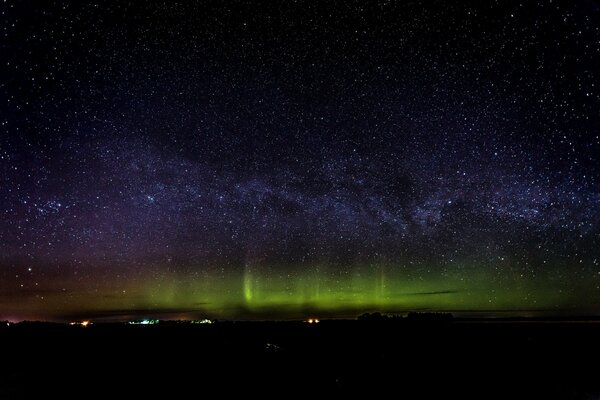 Aurora boreal, tierra oscura, luces de la ciudad y cielo estrellado