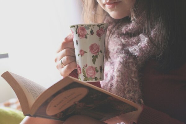 The girl is reading a book and drinking tea from a mug