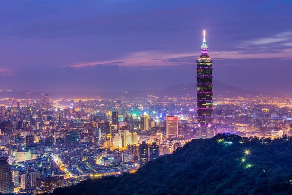 A tall tower in a Chinese city behind green hills in the colors of a purple sunset