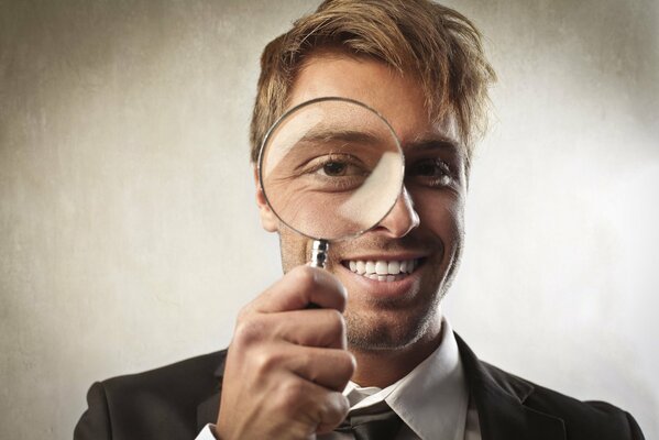 A blond man in a shirt looks through a magnifying glass, smiles