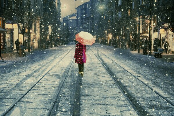 A girl in a red raincoat walks along the tram tracks. sheltering under an umbrella from the wet snow