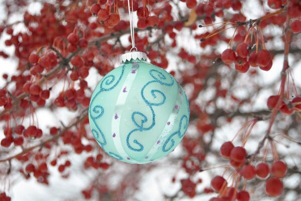 Eine Türkis-Weihnachtskugel mit Mustern hängt im Winter an weißen Fäden auf dem Hintergrund von Zweigen mit kleinen roten Äpfeln