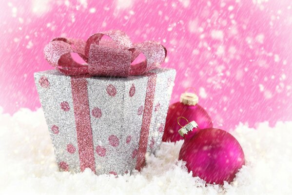 Sulla neve una scatola con un nastro rosa, accanto a due palle di Natale rosa e neve che cade