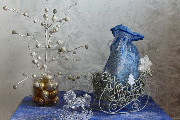Cheval de verre, sac bleu, attaché avec un ruban dans un traîneau, perles blanches, installation d un arbre de Noël dans un pot avec des boules de Noël dorées sur une nappe bleue et blanche sur un fond de mur gris