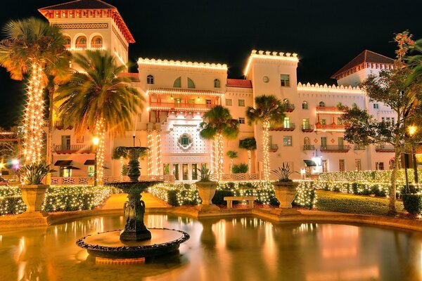 A mansion in lights against the night sky . There are palm trees and a fountain in the courtyard of the mansion