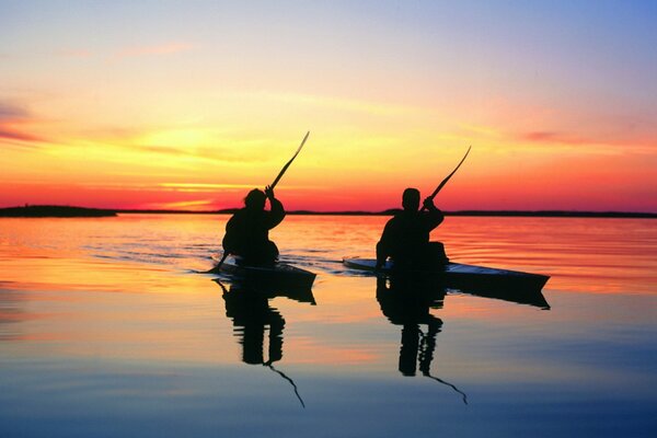 Two people on kayaks float on the water surface at sunset