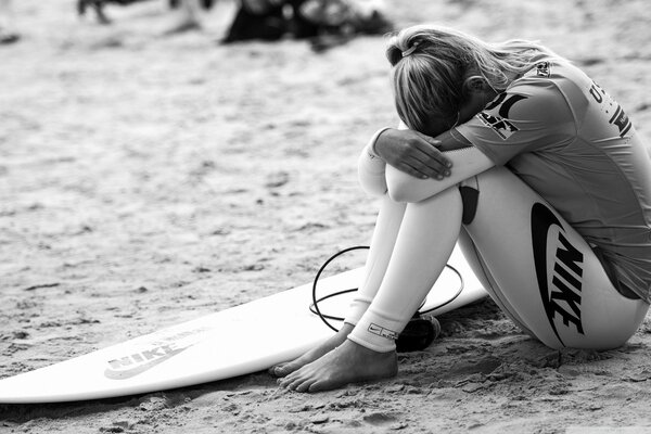 Foto in bianco e nero di una ragazza che seppellisce la testa in ginocchio con una tavola da surf distesa sulla sabbia