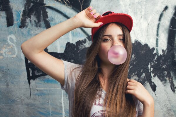 A girl in a red baseball cap inflates a bubble of gum