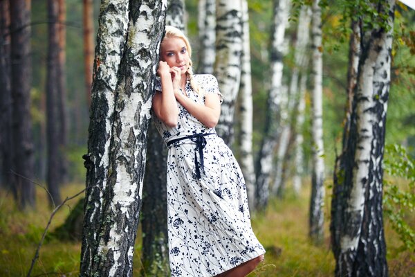 A beautiful blonde girl leaned on a birch tree
