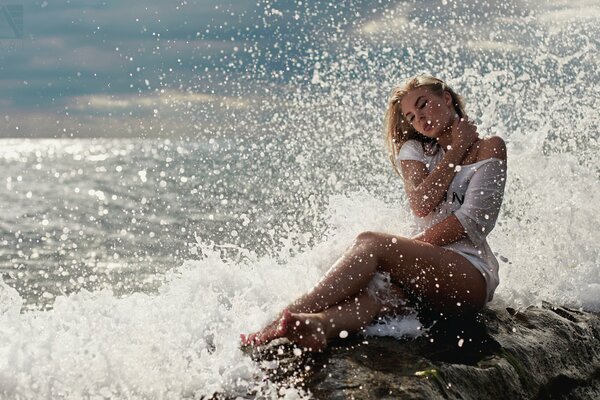 Junge Blondine in weißen Kleidern sitzt am Meer und spritzt