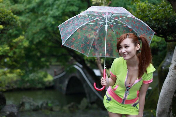 Chica asiática en traje verde claro con escote sonriendo bajo un paraguas transparente en el puente