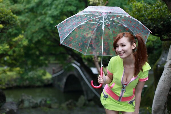 Azjatka w jasnozielonym garniturze z dekoltem uśmiecha się pod przezroczystym parasolem przy moście