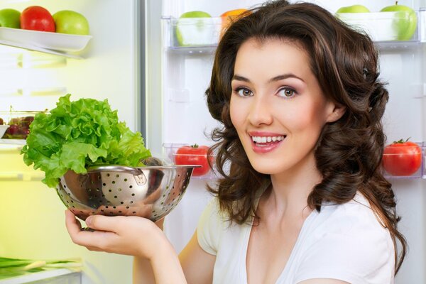 The girl at the refrigerator holds a bowl of salad