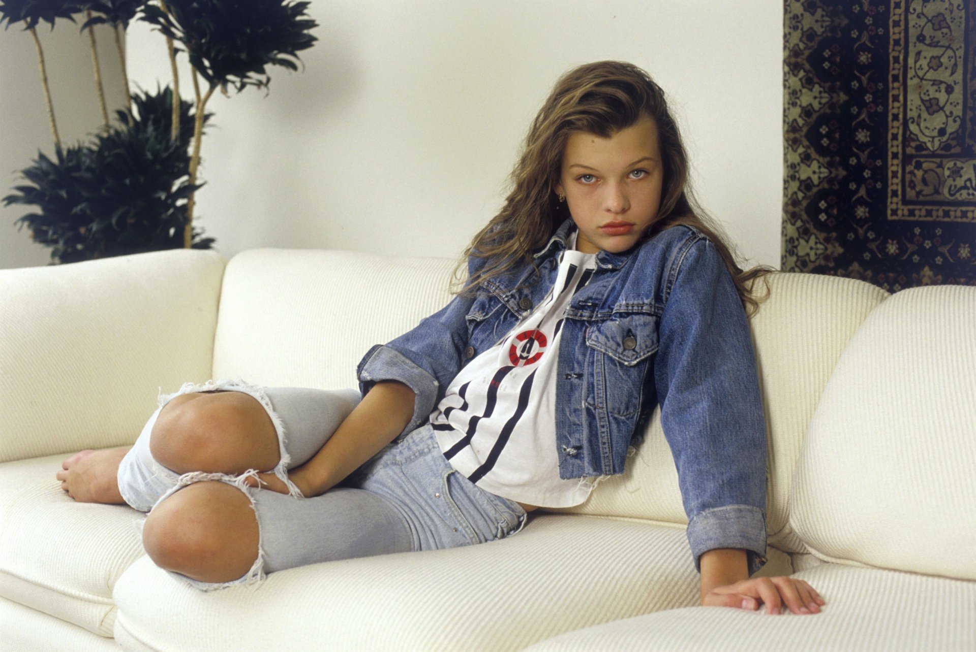 célèbre actrice mila jovovich jeune fille mila en herbe rassurante mila jovovich modèle jovovich look atterrissage hollywood art cinéma film enfant brunette beauté cheveux longs yeux jeans gilet veste canapé pose