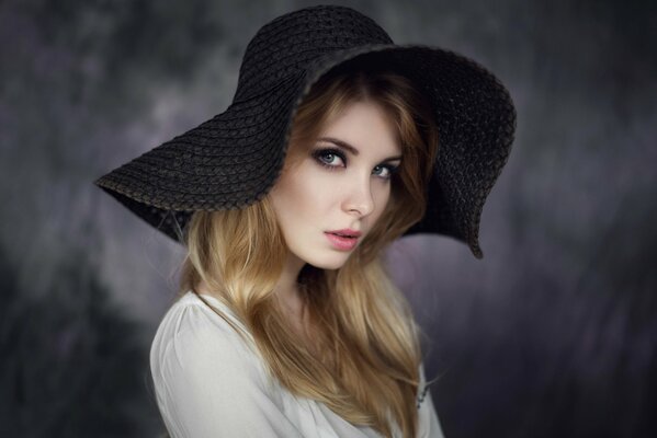 Chica con el pelo suelto y blusa blanca con un gran sombrero negro sobre un fondo negro