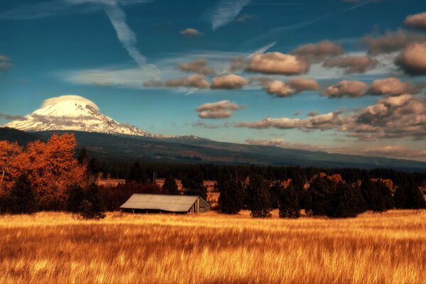 There is a house in the field. Clouds float across the sky hiding behind the horizon and freezing at the mountain