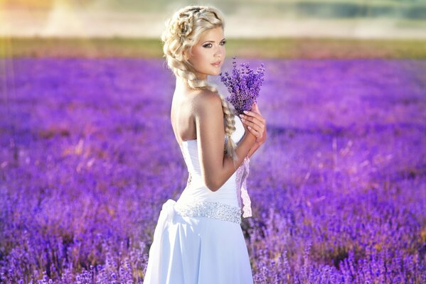 A beautiful girl in a white dress stands in a field with a bouquet of lavender in her hands