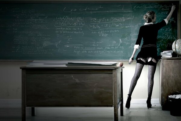 A girl in a black mini with stockings on heels at the blackboard writes with chalk