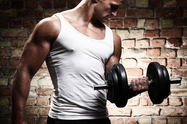 A man in a white T-shirt with dumbbells against a brick wall