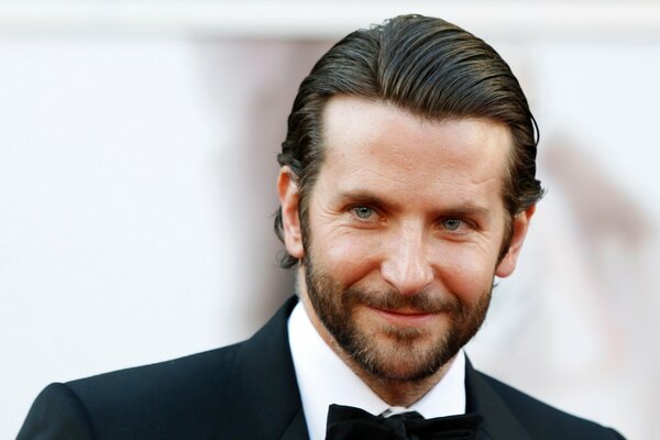 Aktor Bradley Cooper w marynarce i motylu na jasnym tle