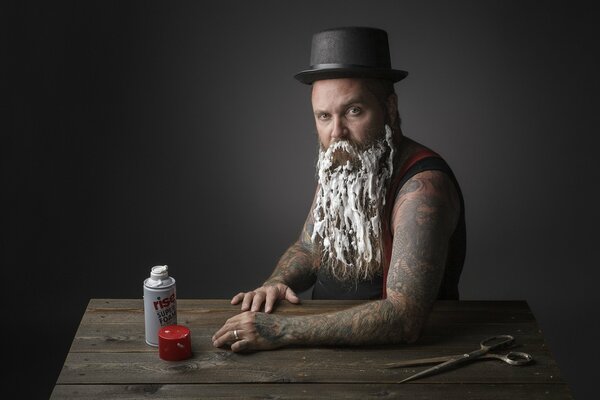 Un homme avec un chapeau et des tatouages barbouillé de mousse longue barbe