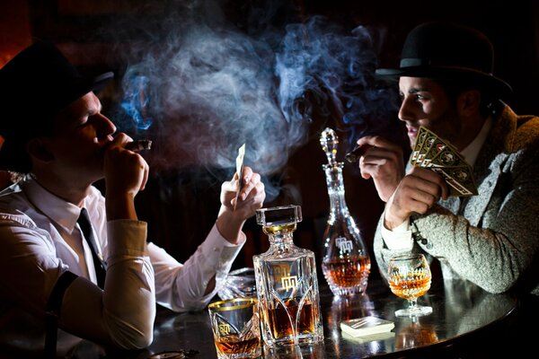 Двое мужчин играют в карты, курят и выпивают за столом