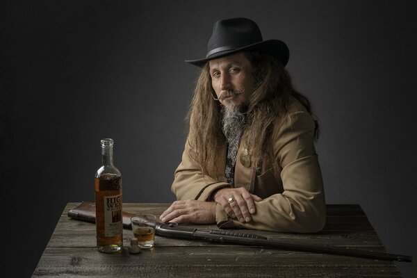 A long-haired man with a beard and a hat in the image of a cowboy is sitting at a wooden table, on which there is a bottle of whiskey and a half-filled glass, as well as a gun