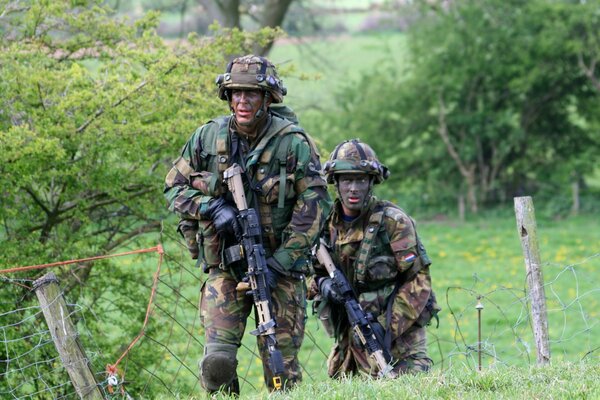 Dwóch żołnierzy z bronią porusza się po zielonym polu wśród drzew