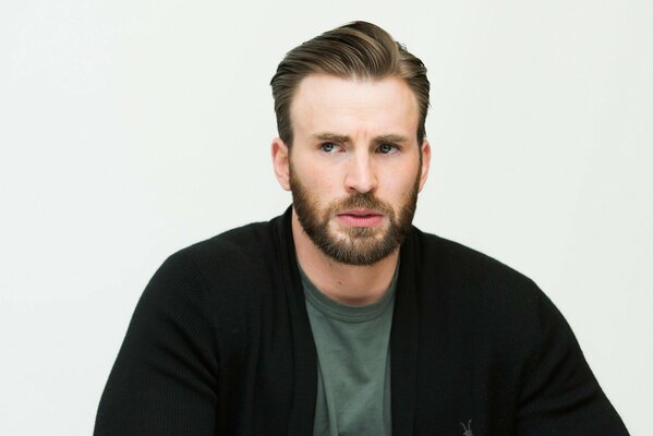 Actor Chris Evan on a white background with stubble in a black jacket and a green T-shirt looks intently to the right