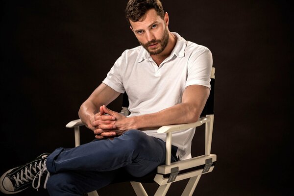 Acteur, mannequin et musicien Jamie Dornan dans un t-shirt blanc, un pantalon bleu et des baskets noires est assis dans une chaise en bois, les doigts croisés et jetant la jambe gauche sur le genou droit sur un fond noir