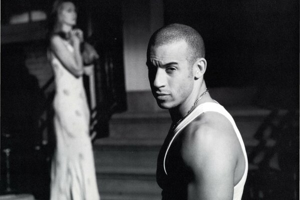 Vin Diesel is standing in profile and looking into the camera with a woman standing in the background