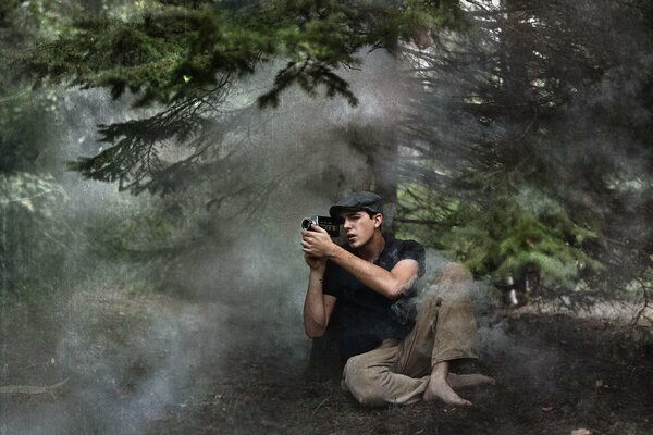 Giovane fotografo seduto a piedi nudi a terra in una foresta di conifere nebbiosa e fotografando