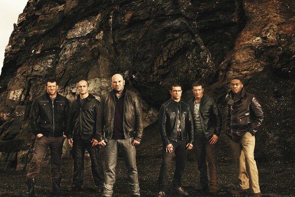 Six men in leather jackets are standing near a rock