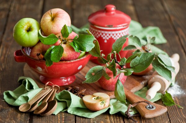 Sulla tavola di legno frutta in fruttiera, mattarello, casseruola, cucchiai di legno e mele con foglie