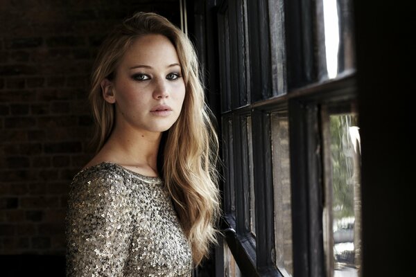 A girl in a shiny dress and loose hair is standing by the window