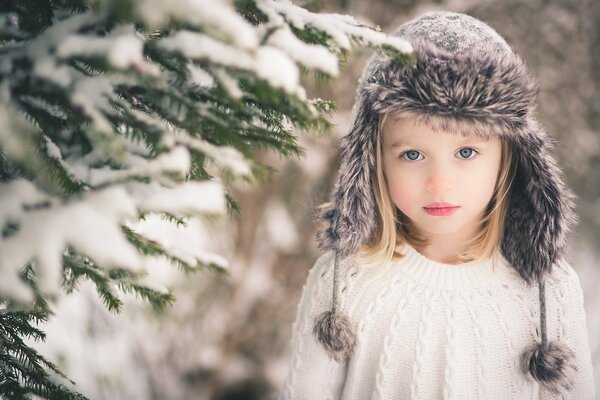 Niña de ojos grises con sombrero en un árbol de Navidad cubierto de nieve