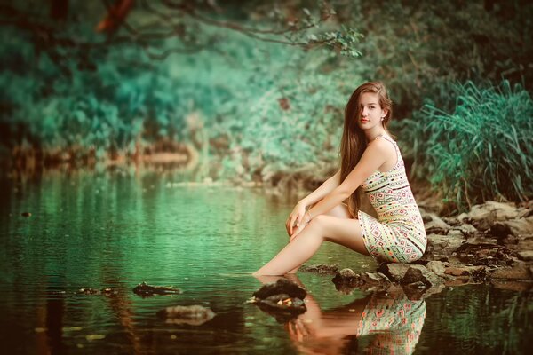The girl is sitting in the forest on the rocks next to the river