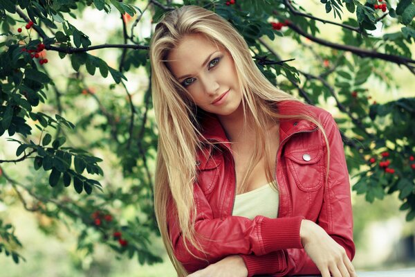 A beautiful blonde girl in a red jacket with a mesmerizing look against the background of tree branches with green foliage and red berries