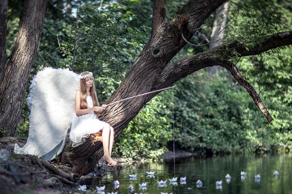 Jeune fille à l image d un ange avec des ailes blanches assis sur un arbre avec une canne à pêche sur un paysage d été de l étang