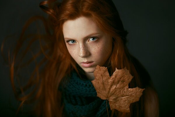 A red-haired girl with freckles in a knitted green scarf holds a dry orange maple leaf