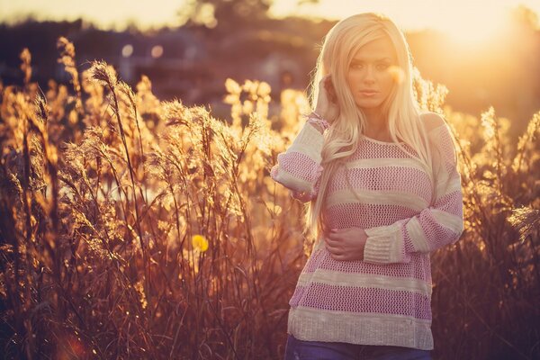 A blonde in a pink and white striped jacket stands against the background of tall grass and the setting sun
