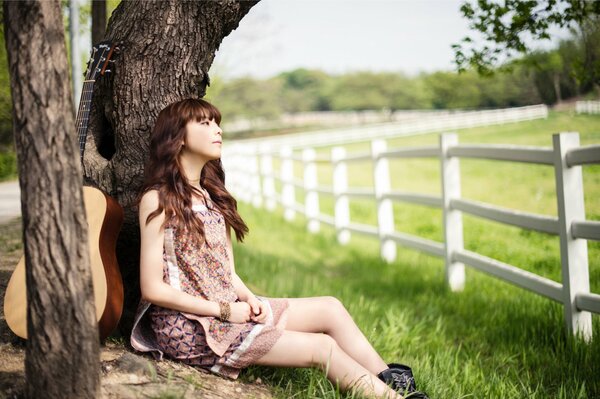 A girl with a guitar sits leaning on a tree and looks into the distance