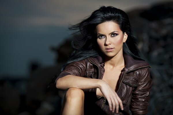 A burning brown-eyed brunette in a brown leather jacket sits against the background of evening rocky nature