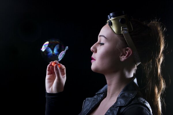 A girl in motorcycle glasses looks at a soap bubble in which a blue butterfly is enclosed