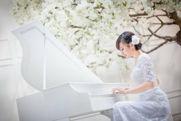 Chica asiática en un vestido blanco toca un piano blanco sobre un fondo blanco