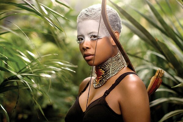 Una mujer de piel oscura con una cabeza de combate con un collar étnico y un arco de batalla en su hombro se encuentra en medio de la jungla