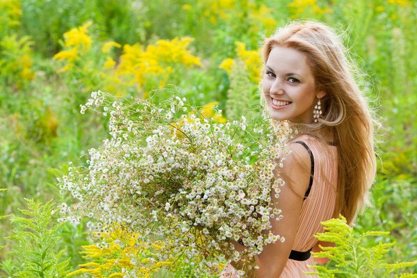 Ein lächelndes Mädchen in langen Ohrringen steht mit einem Strauß weißer Wildblumen in einem rosa Kleid inmitten des Grüns