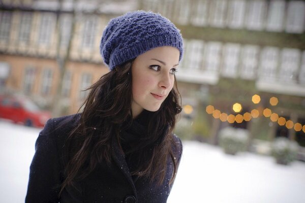 A brown-haired woman in a purple hat and a purple coat stands against the background of a winter city with Christmas lights